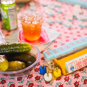 May include: A clear glass bowl filled with green and brown dill pickles on a pink and white floral tablecloth. A glass of iced tea, a can of soda, and a set of mahjong tiles are also visible on the table.