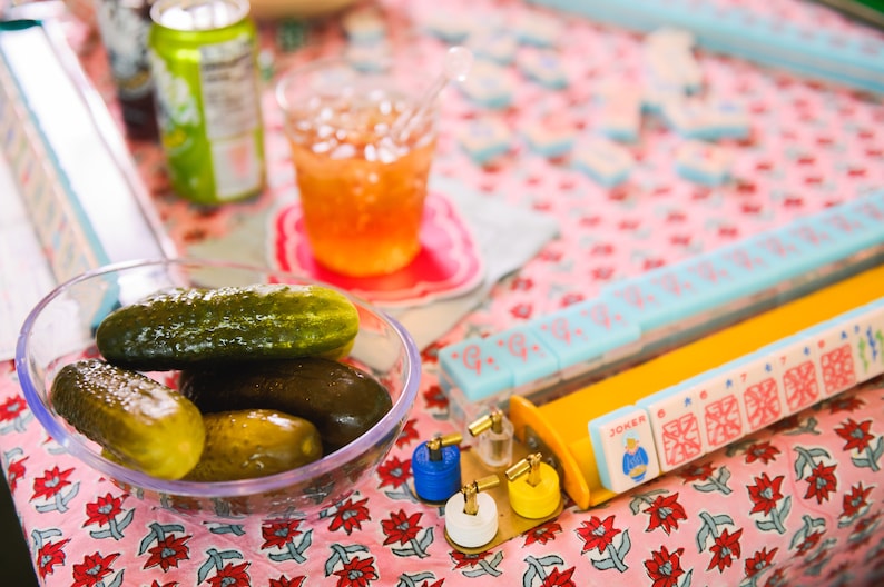 May include: A clear glass bowl filled with green and brown dill pickles on a pink and white floral tablecloth. A glass of iced tea, a can of soda, and a set of mahjong tiles are also visible on the table.