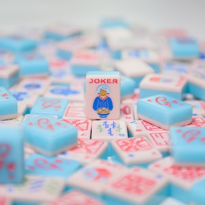 Op de afbeelding: Een close-up van een stapel blauwe en witte mahjong-tegels met een enkele joker-tegel in het midden. De joker-tegel toont een cartoonvrouw in een blauwe jurk en een rode hoed.