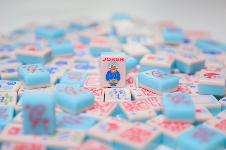 May include: A close-up of a pile of blue and white mahjong tiles with a single joker tile in the center. The joker tile features a cartoon woman in a blue dress and a red hat.