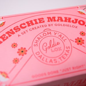 May include: Pink box with the text "MENSCHIE MAHJONG" in red letters. The box also has a circular sticker with the text "SHALOM Y'ALL Goldie LOX DALLAS, TEXAS" in pink letters.