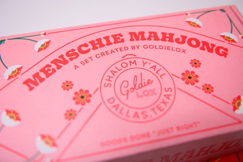 May include: Pink box with the text "MENSCHIE MAHJONG" in red letters. The box also has a circular sticker with the text "SHALOM Y'ALL Goldie LOX DALLAS, TEXAS" in pink letters.