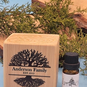 May include: A wooden block engraved with a tree design and the text "Anderson Family EST.2013". Next to it is a small brown glass bottle with a black cap and a label that reads "Cedar Oil". The background includes greenery.
