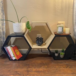 May include: A set of three hexagonal shelves in black and natural wood finishes. The shelves are stacked on top of each other and feature a variety of items, including books, plants, and a clock.