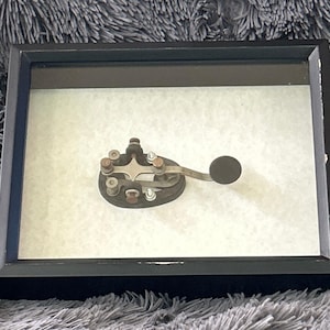 May include: A vintage telegraph key mounted inside a rectangular black shadow box frame. The telegraph key features a dark base, metallic components, and a circular knob. The frame has a glass front and a light-colored backing behind the key.