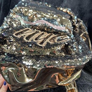 Peut inclure: Un sac à dos à paillettes noir et or avec le mot "NYC" brodé en fil d'or. Le sac à dos a un fond métallique doré et une fermeture éclair rose. Le sac est conçu avec une surface brillante et réfléchissante.