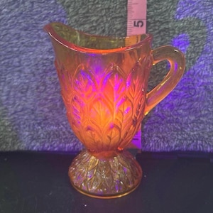 May include: An amber-colored glass creamer with a handle and a decorative leaf pattern. The creamer is approximately 5 inches tall, with a pedestal base and a flared top. The glass has a warm, glowing appearance.