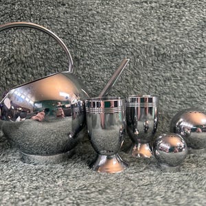 May include: A set of polished silver serving pieces, including a watering can-shaped pitcher with a handle, two egg cups, and two spherical salt and pepper shakers. The items are reflective, with a shiny surface.
