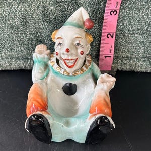 May include: A vintage ceramic clown figurine with a smiling face, red nose, and a pointed hat with a red pom-pom. The clown is wearing a light blue and orange outfit with a gold-colored collar. The figurine is approximately 3 inches tall.
