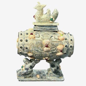 May include: An antique, beige-colored decorative urn with a bird-like figure on top. The urn is supported by two figures and adorned with small, colorful stones. The design features intricate carvings and a weathered appearance, suggesting an aged artifact.