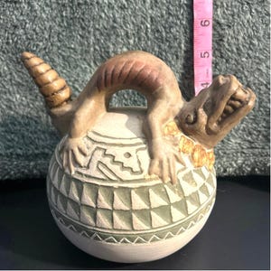 May include: A ceramic vessel with a lizard-like creature perched on top. The vessel is off-white with green and brown geometric patterns. The lizard is tan with brown and gold accents. The pink measuring tape shows the vessel is approximately 5 inches tall.