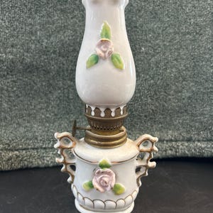 May include: A small, white ceramic oil lamp with a decorative floral design. The lamp features a pink rose and green leaves, with gold-colored accents. The lamp has a brass-colored oil reservoir and a glass chimney.