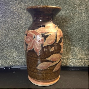 May include: A brown ceramic vase with a flared top and a wide base. The vase features a leaf design in shades of peach and brown. The vase is approximately 7 inches tall.