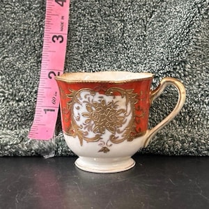 May include: A small antique teacup with a white base and a red rim. The cup features gold floral designs and a gold handle. The cup is approximately 2 inches tall.