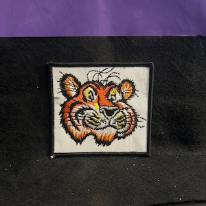 May include: A square embroidered patch featuring a stylized tiger face with orange, yellow, and black thread on a white background. The tiger has a wide grin, prominent whiskers, and a black stitched border framing the entire square design.