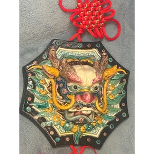 May include: A ceramic dragon face wall hanging with a black octagonal frame. The dragon has a detailed face with red, blue, yellow, and green accents. A red decorative knot hangs above the dragon.