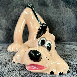 May include: A ceramic figurine of a cartoon dog, lying down with its legs in the air. The dog has a light tan body, black ears, a black nose, and a red mouth. The figurine is glossy and appears to be hand-painted.