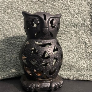 May include: A black owl-shaped candle holder with cut-out designs. The owl has large eyes and a textured surface. The base features clawed feet. This decorative item is suitable for Halloween or other seasonal decor.