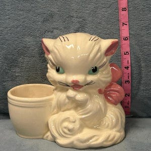 Peut inclure: Un pot de fleurs vintage en céramique crème avec des accents roses. Le chat a des yeux verts, un nœud rose et un petit pot de fleurs attaché. Le pot mesure environ 7.6 cm de haut.