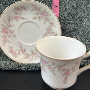 May include: A white teacup and saucer set with a delicate pink rose pattern and gold trim. The cup has a curved handle and the saucer is round. The floral design is repeated on both pieces, creating a cohesive look.