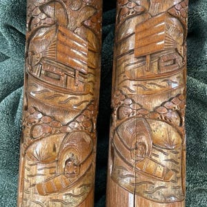 May include: Two cylindrical brown wooden carvings featuring detailed relief scenes of traditional houses, trees, and small boats on water. The wood displays natural grain patterns and vertical cracks, with intricate hand-carved textures throughout.