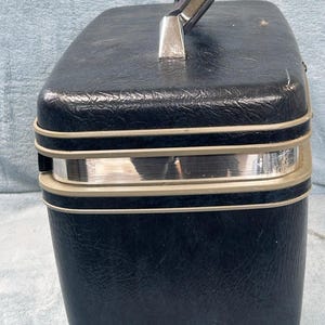 May include: A vintage black vanity case with a textured exterior. The case features a silver handle and trim, with a rectangular shape. The handle is attached with silver metal hardware. The case is designed for travel and storage.