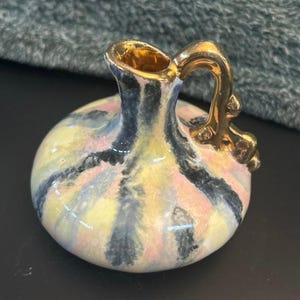 May include: A small, decorative ceramic vase with a round body and a gold handle and rim. The vase is painted with a colorful pattern of yellow, pink, and black stripes. The handle has a decorative design.