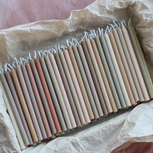 May include: A selection of thin, pastel-coloured candles arranged in a rectangular formation, presented on a bed of crumpled, light-brown paper. The candles display a range of beige, pink, and green hues. The backdrop is a soft, pink fabric.