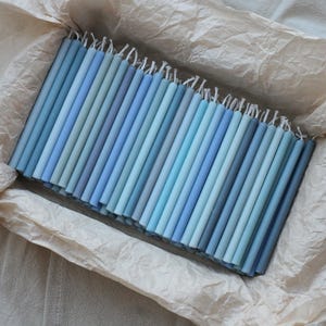 Peut inclure: Un assortiment de bougies coniques bleues de diff&eacute;rentes nuances, dispos&eacute;es dans une bo&icirc;te doubl&eacute;e de papier. Les bougies varient du bleu clair au bleu fonc&eacute;, avec des m&egrave;ches blanches visibles en haut. L'ensemble d&eacute;gage une atmosph&egrave;re douce et apaisante.