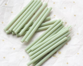 May include: A bundle of thin, sage green taper candles with white wicks. The candles are arranged in two rows on a white surface with gold polka dots.