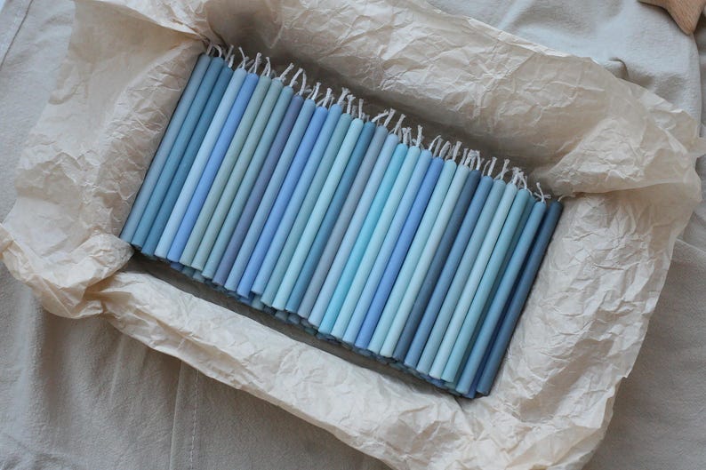 Peut inclure: Un assortiment de bougies coniques bleues de diff&eacute;rentes nuances, dispos&eacute;es dans une bo&icirc;te doubl&eacute;e de papier. Les bougies varient du bleu clair au bleu fonc&eacute;, avec des m&egrave;ches blanches visibles en haut. L'ensemble d&eacute;gage une atmosph&egrave;re douce et apaisante.