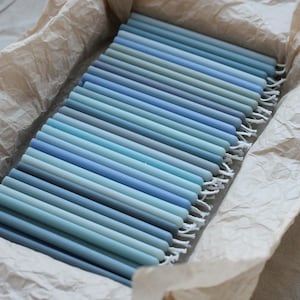 Peut inclure: Un assortiment de bougies coniques bleues de diff&eacute;rentes nuances, dispos&eacute;es dans un r&eacute;cipient rectangulaire doubl&eacute; de papier beige. Les bougies varient du bleu clair au bleu fonc&eacute;, cr&eacute;ant un effet de d&eacute;grad&eacute;. Le r&eacute;cipient est pos&eacute; sur un tissu de couleur neutre.