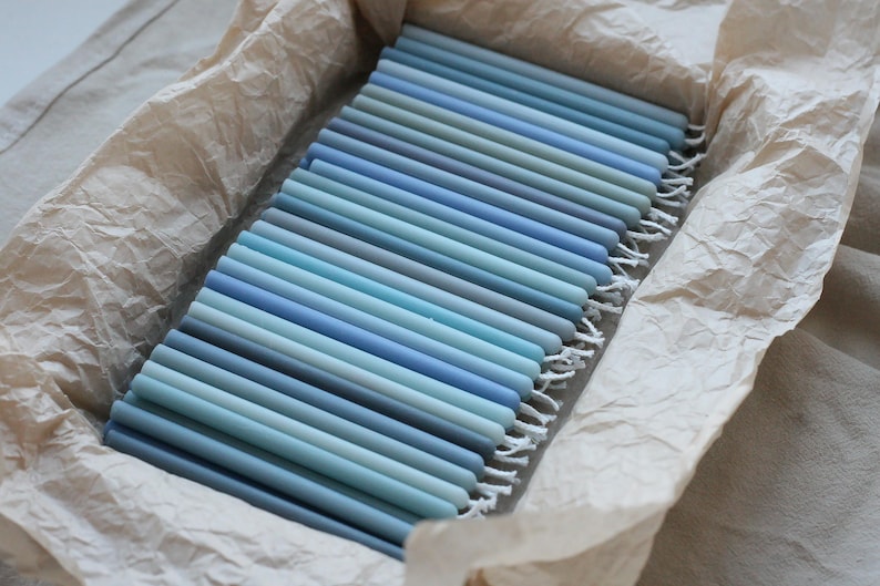 Peut inclure: Un assortiment de bougies coniques bleues de diff&eacute;rentes nuances, dispos&eacute;es dans un r&eacute;cipient rectangulaire doubl&eacute; de papier beige. Les bougies varient du bleu clair au bleu fonc&eacute;, cr&eacute;ant un effet de d&eacute;grad&eacute;. Le r&eacute;cipient est pos&eacute; sur un tissu de couleur neutre.