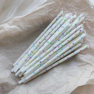 Puede incluir: Un conjunto de velas de cumpleaños blancas con lunares de colores. Las velas están dispuestas sobre una superficie de papel arrugado de color marrón claro. Cada vela es larga y delgada, con una pequeña mecha en la parte superior.