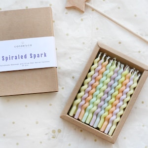 Può includere: Un set di candele a spirale dai colori pastello in una scatola rettangolare. La scatola è aperta, rivelando le candele disposte in file. Una scatola chiusa con la scritta "Spiraled Spark" è accanto alla scatola aperta. Anche una bacchetta a stella di legno è nell'immagine.