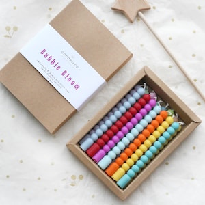 Puede incluir: Un juego de velas de burbujas coloridas en una caja de cartón, con el texto "Bubble Bloom" en la etiqueta. Las velas están dispuestas en filas de varios colores. También hay una varita de estrella de madera.