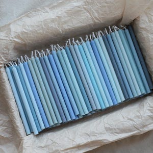 Peut inclure: Un ensemble de bougies coniques bleues de diff&eacute;rentes nuances, dispos&eacute;es dans un contenant doubl&eacute; de papier. Les bougies, allant du bleu clair au bleu fonc&eacute;, cr&eacute;ent un effet de d&eacute;grad&eacute;. Les m&egrave;ches sont visibles en haut.