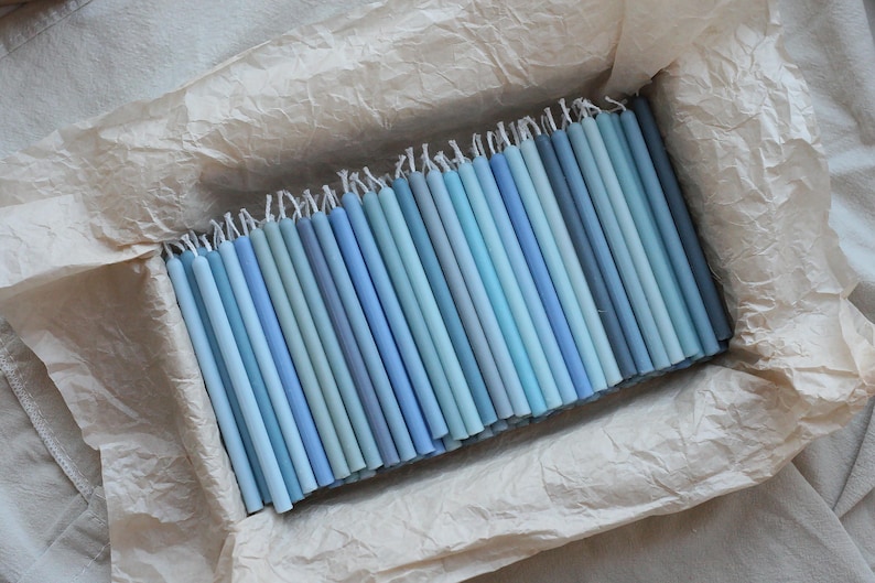 Peut inclure: Un ensemble de bougies coniques bleues de diff&eacute;rentes nuances, dispos&eacute;es dans un contenant doubl&eacute; de papier. Les bougies, allant du bleu clair au bleu fonc&eacute;, cr&eacute;ent un effet de d&eacute;grad&eacute;. Les m&egrave;ches sont visibles en haut.