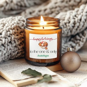 Könnte beinhalten: Eine brennende, bernsteinfarbene Kerze in einem Glas mit einem weißen Etikett mit der Aufschrift "happy holidays to the one & only". Eine goldene Kugel liegt neben der Kerze. Die Kerze steht auf einem Buch mit einer Strickdecke im Hintergrund.
