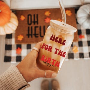 Könnte beinhalten: Ein durchsichtiger Glasbecher mit Holzdeckel und Glasstrohhalm. Der Becher ist mit einem hellfarbigen Getränk gefüllt und trägt den Text "HERE FOR THE BIRTH" in roter und rosa Schrift. Der Becher wird in einer Hand gehalten.