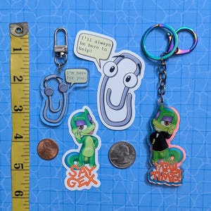 May include: Set of four keychains featuring a green lizard character. One keychain has the text "Say Gex" and another has the text "Tail Time!" The other two keychains are a paperclip with googly eyes and a paperclip with headphones.