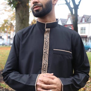 Puede incluir: Una camisa negra de manga larga con ribete beige y un patrón geométrico en la parte delantera. La camisa tiene un cuello mandarín y un bolsillo en el lado izquierdo.