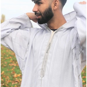 Op de afbeelding: Een witte gekapte robe met een zilveren geborduurde rand. De robe heeft een gestreept patroon en wordt gedragen door een persoon met krullend haar en een baard.