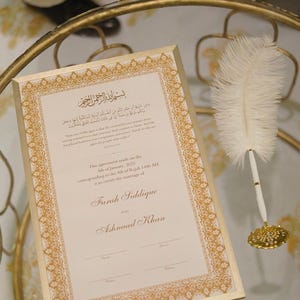 Puede incluir: Un certificado enmarcado en oro con una pluma de pluma blanca y un tintero. El certificado es para el matrimonio de Farah Siddique y Ashnaad Khan. La fecha del matrimonio es el 4 de enero de 2025, correspondiente al 4 de Rajab 1446 AH.