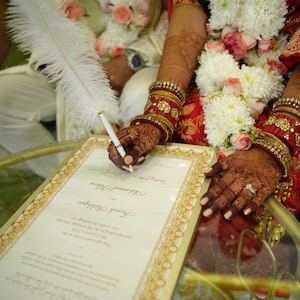 Puede incluir: Una novia y un novio firmando un certificado de matrimonio con un bol&iacute;grafo de pluma blanca. La novia lleva un atuendo tradicional indio con joyas rojas y doradas. El novio lleva un sherwani blanco con una guirnalda de flores alrededor del cuello.