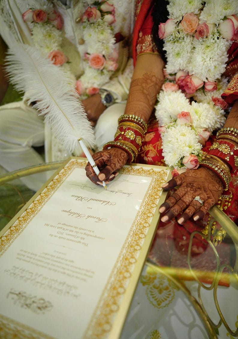 Puede incluir: Una novia y un novio firmando un certificado de matrimonio con un bol&iacute;grafo de pluma blanca. La novia lleva un atuendo tradicional indio con joyas rojas y doradas. El novio lleva un sherwani blanco con una guirnalda de flores alrededor del cuello.