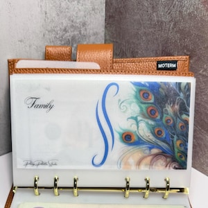 May include: A brown leather bound planner with a clear plastic pocket that has a peacock feather design and the word "Family" written on it. The planner has a black label that says "MOTERM".