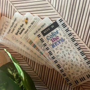 May include: A set of five printable coloring pages for kids featuring a piano keyboard with musical notes and the text "Color the Keys". The pages are printed on transparent paper and are designed for use with markers or crayons.