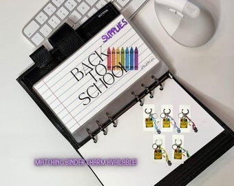 May include: A black leather binder with a lined paper insert that reads "BACK TO SCHOOL" with colourful crayon illustrations. The word "SUPPLIES" is written above the text. Several keychain charms are also visible.