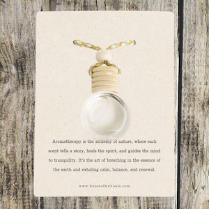 May include: A white card with a quote about aromatherapy. The quote reads: "Aromatherapy is the alchemy of nature, where each scent tells a story, heals the spirit, and guides the mind to tranquility. It's the art of breathing in the essence of the earth and exhaling calm, balance, and renewal." The card also includes the website address: www.houseofwrituals.com. A small glass bottle with a wooden cap and a string is hanging from the top of the card.